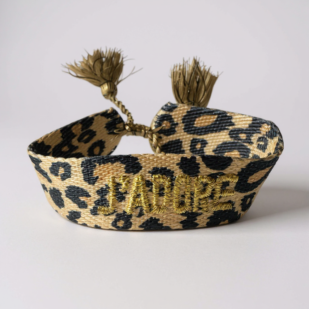Bracelet J’ADORE léopard en tissu tissé avec broderie dorée et pompons ajustables.